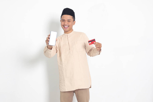 Retrato del atractivo hombre musulmán asiático en camisa koko con gorra de calavera sosteniendo una tarjeta de crédito mientras muestra la pantalla vacía en el teléfono móvil para maqueta y gráfico. Imagen aislada sobre fondo blanco - Foto, imagen