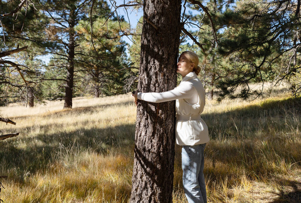 Mujer rubia en tela casual abraza un árbol en el bosque o el parque, disfrutando de su sombra. Día Nacional del Amor a los Árboles, 16 de mayo. Copiar espacio para texto. Cuidando de la Naturaleza, Wood. Oxitocina, sentimientos de bienestar - Foto, Imagen