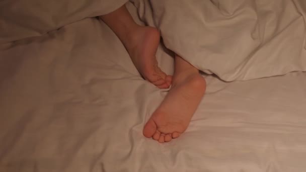 los pies inquietos de un niño en una sábana debajo de una manta por la noche. Bebé niña con las piernas desnudas lanza y no se duerme en la cama en el dormitorio por la noche - Metraje, vídeo