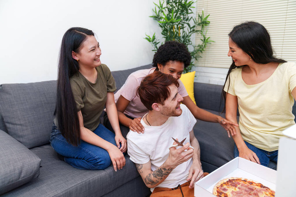 Χαρούμενη ποικιλόμορφη ομάδα φίλων μοιράζονται ένα γέλιο με πίτσα σε έναν καναπέ. Ζωηρή αλληλεπίδραση σε ένα σύγχρονο σαλόνι. - Φωτογραφία, εικόνα