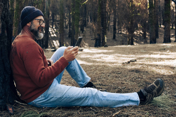 Один человек пользуется телефоном в лесу, наслаждаясь технологиями и активным отдыхом на свежем воздухе. Современный человек говорит на клеточном уровне с деревьями на заднем плане. Люди и общение о путешествиях - Фото, изображение