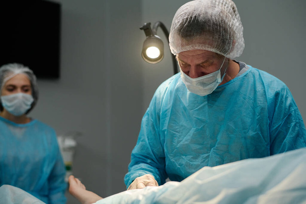 Ammatillinen kirurgi lääketieteellisissä pensaikoissa seisoo leikkauspöydällä potilaan kanssa, joka on peitetty lautasliinalla kirurgisen toimenpiteen aikana - Valokuva, kuva