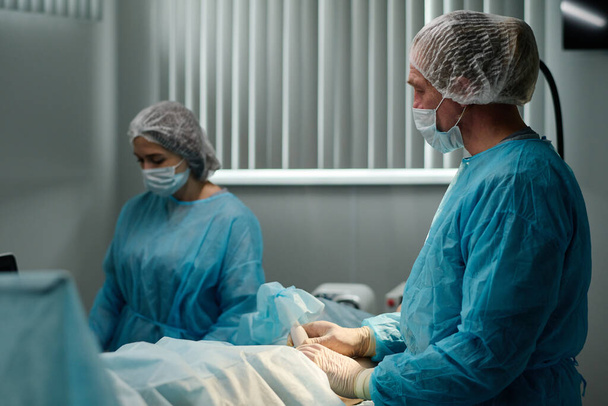 Erfahrener Arzt in Schutzmaske, Handschuhen und medizinischem Peeling mit Sonde bei chirurgischen Eingriffen am Bein oder Knie des Patienten - Foto, Bild