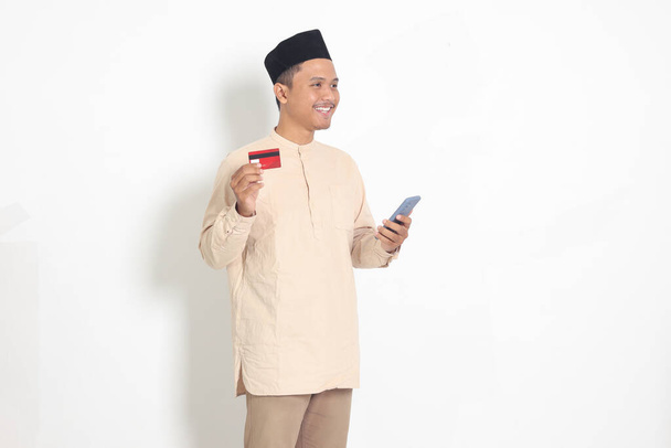 Retrato del atractivo hombre musulmán asiático en camisa koko con gorra de calavera sosteniendo un teléfono móvil y presentando tarjeta de crédito. Imagen aislada sobre fondo blanco - Foto, imagen