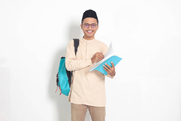 Portrait of excited student Asian muslim man in koko shirt with skullcap carrying backpack, while holding his school books. Islamic education concept. Isolated image on white background - Photo, Image