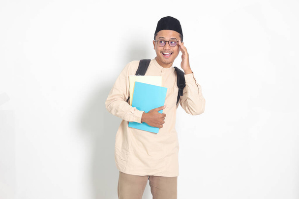 Portrait d'étudiant asiatique musulman excité en chemise koko avec calotte portant un sac à dos, tout en tenant ses livres scolaires. Concept d'éducation islamique. Image isolée sur fond blanc - Photo, image