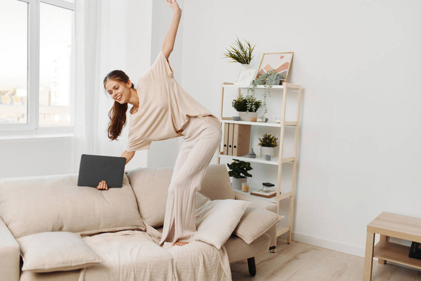 Усміхнена жінка працює на ноутбуці в затишній вітальні, насолоджуючись позаштатною роботою в сучасній домашній обстановці - Фото, зображення