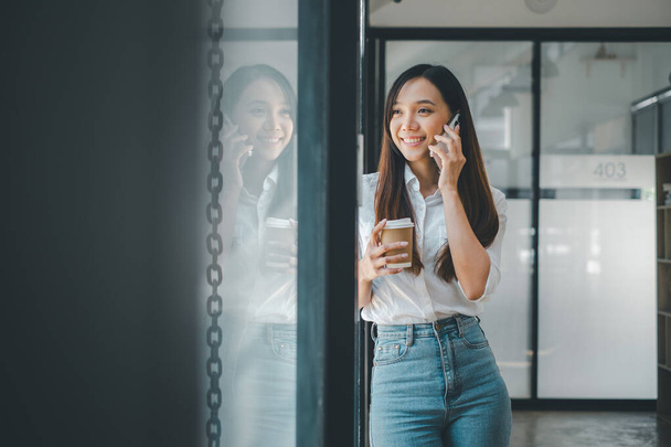 Business concetto di tecnologia, Una donna professionale prende una telefonata mentre tiene in mano una tazza di caffè, in piedi vicino alla finestra in un corridoio di ufficio illuminato dal sole - Foto, immagini