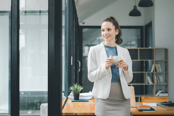 Концепція ділової жінки: усміхнена бізнес-леді насолоджується кавовою перервою, стоячи розслабленою в стильному і сучасному офісі - Фото, зображення