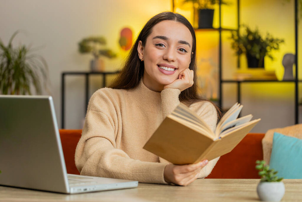 Καυκάσια χαμογελαστή γυναίκα που διαβάζει ενδιαφέρον βιβλίο, γυρίζει σελίδες χαμογελώντας απολαμβάνοντας τη λογοτεχνία, ξεκουράζεται στο τραπέζι. Πορτρέτο της ειρηνικής χαρούμενο κορίτσι χαλαρώνοντας στο σπίτι διαμέρισμα σαλόνι - Φωτογραφία, εικόνα