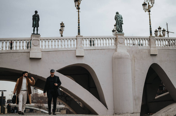 Δύο κομψοί άντρες συζητούν ενώ περπατούν κάτω από μια γέφυρα στολισμένη με λάμπες και αγάλματα. Πόλη ατμόσφαιρα ζωή και την έννοια της φιλίας. - Φωτογραφία, εικόνα