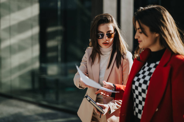 A pair of young businesswomen engaging in a discussion over documents on a sunny day, exuding confidence and teamwork outside an office building. - Photo, Image