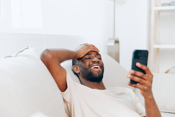 Hombre afroamericano feliz usando Smartphone y relajarse en el sofá negro en casa - Foto, imagen