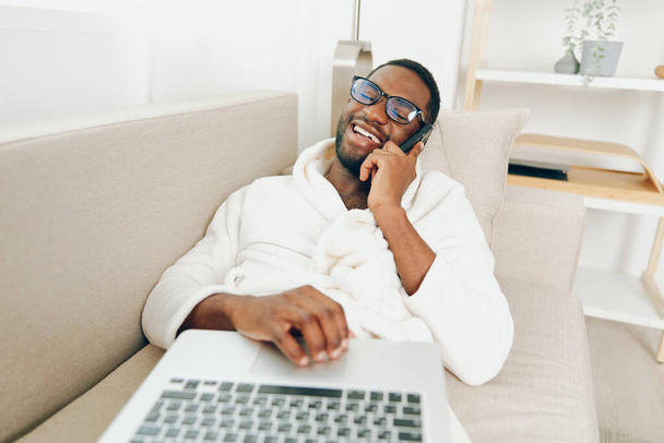 Gülümseyen Afrikalı Amerikalı Adam rahat bir oturma odasında dizüstü bilgisayarda çalışıyor. Görüntü, neşeli ve odaklı bir serbest çalışanı, bornoz giymiş, dizüstü bilgisayarlı bir koltukta oturuyor. - Fotoğraf, Görsel