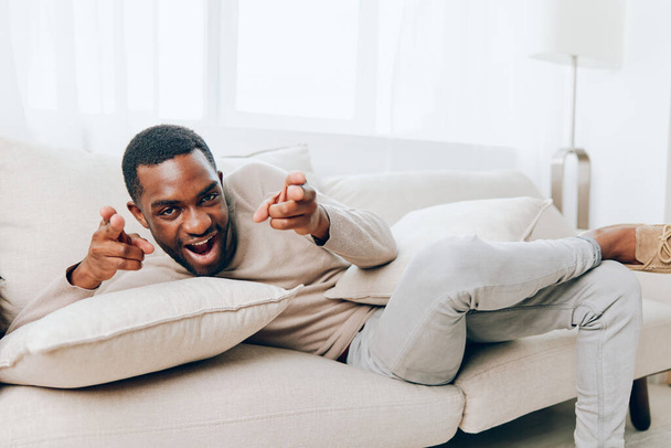 Pihentetett afro-amerikai férfi ül egy kényelmes kanapén a nappaliban, mosolygott és tartott egy párnát Élvezi a békés hétvégét otthon, néz valamit online és gondolkodás egy - Fotó, kép