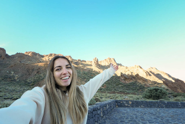 Uma jovem está capturando um momento alegre como ela toma uma selfie com as montanhas pitorescas banhadas no brilho suave do sol da noite no fundo. - Foto, Imagem