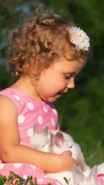 Щасливий малюк одягнений в рожеве плаття з посмішкою, тримає білого кролика, оточеного людьми на природі, з травою і насолоджується дозвіллям і веселощами. - Кадри, відео