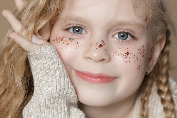 Portret van een schattig blond meisje met blauwe ogen en glitters op haar gezicht. - Foto, afbeelding
