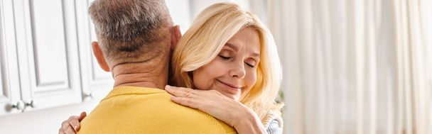 A mature loving couple in cozy homewear, the woman embracing the man in a yellow sweater in a warm, affectionate hug. - Photo, Image