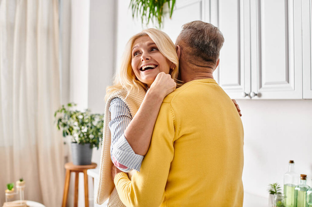 A mature loving couple in cozy homewear sharing a warm hug in the kitchen of their home. - Photo, Image