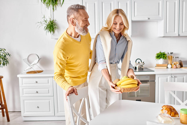 Un uomo e una donna maturi stanno nella loro accogliente cucina, tenendo in mano le banane e condividendo un dolce momento insieme. - Foto, immagini