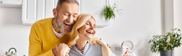 Ein reifer Mann und eine reife Frau in Hauskleidung teilen einen freudigen Moment, als sie gemeinsam in einer gemütlichen Küche zu Hause lachen. - Foto, Bild