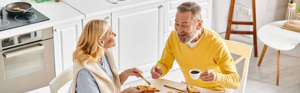 A mature loving couple enjoying a meal together at a table in cozy homewear, savoring delicious food in a warm atmosphere. - Photo, Image
