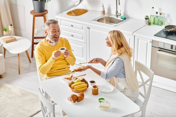 Una pareja madura y cariñosa en ropa de casa acogedora sentados juntos en una mesa de cocina, disfrutando de un momento de unión en casa. - Foto, Imagen