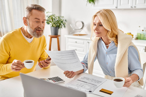 A mature man and woman in comfy attire sit at a table, engaged in reviewing and discussing paperwork together. - Photo, Image