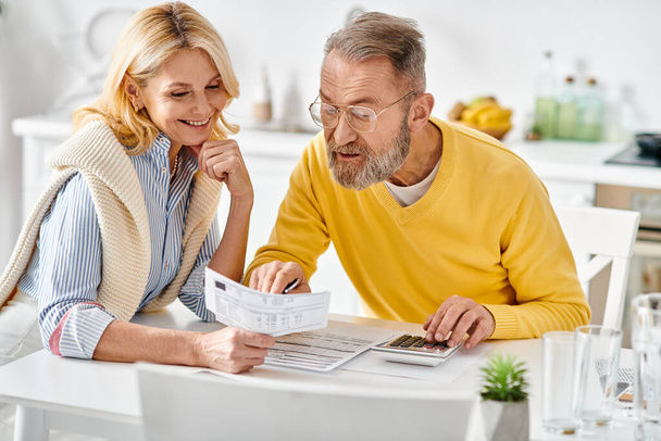 A mature man and woman in cozy homewear are seated in the kitchen, engrossed in reading the newspaper together. - Photo, Image