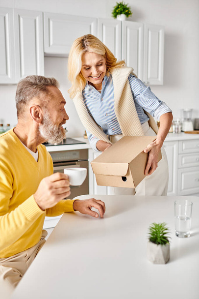 Dojrzały mężczyzna i kobieta w przytulnym homewear otwierają pudełko na blacie kuchennym, ciekawi i podekscytowani jego zawartością. - Zdjęcie, obraz