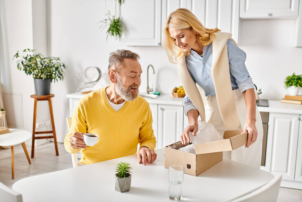A mature loving couple in cozy homewear, opening a box to reveal a plant inside, sharing a moment of joy and discovery in their kitchen. - Photo, Image