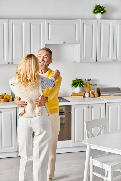 Een volwassen liefdevol paar in gezellige huiskledij dansen samen in de keuken, genieten van een moment van verbinding en vreugde. - Foto, afbeelding