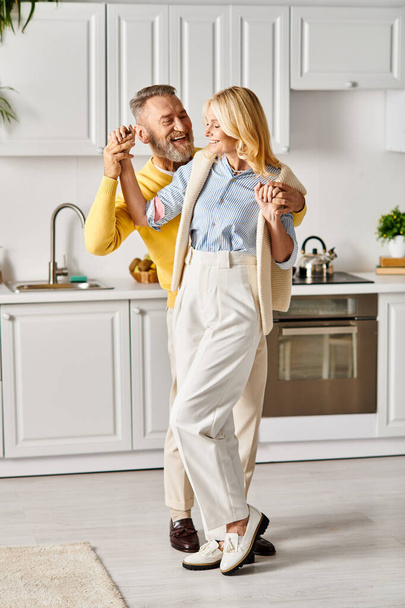 Un couple aimant mature dans des vêtements confortables dansant joyeusement dans leur cuisine, se connectant par le mouvement et la musique. - Photo, image