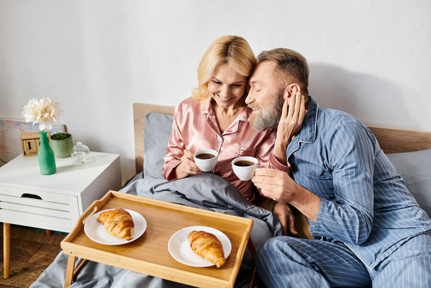 Ένα ώριμο ζευγάρι στο ζεστό homewear απολαμβάνοντας ένα χαλαρωτικό πρωινό στο κρεβάτι τους με καφέ και γλυκά. - Φωτογραφία, εικόνα