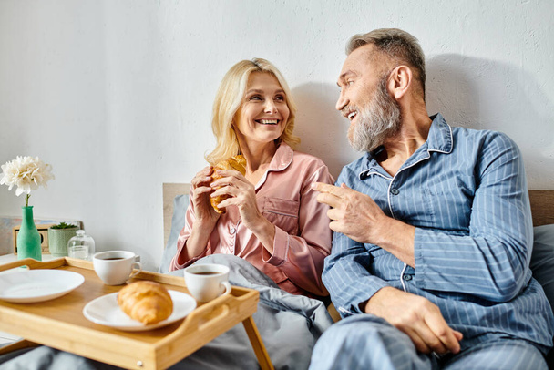 A mature loving couple relaxes on a couch, enjoying a meal together in their bedroom. - Photo, Image