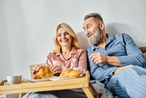 Una pareja madura y cariñosa en ropa de casa sentada en un sofá, disfrutando de una bandeja de croissants juntos en un ambiente acogedor. - Foto, Imagen