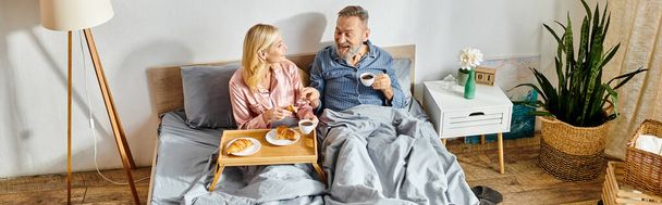 A mature loving couple, dressed in cozy homewear, sit closely together on a bed, sharing a moment of tenderness and connection. - Photo, Image