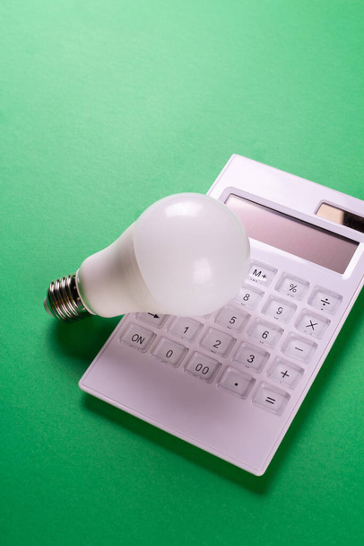 Білий калькулятор і світлодіодні лампи на зеленому фоні. Концепція, що показує оплату рахунків за електроенергію. Концепція економії електроенергії. Зниження оплати комунальних платежів - Фото, зображення
