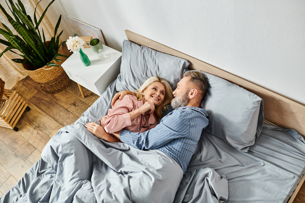 Een volwassen liefdevol paar in huiskledij knuffelen en ontspannen samen op een bed, het delen van een rustig moment van intimiteit. - Foto, afbeelding