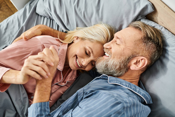 居心地の良いホームウェアの成熟した愛するカップルは,ベッドに平和的に横たわり,彼らの寝室で柔らかい抱擁を共有します. - 写真・画像