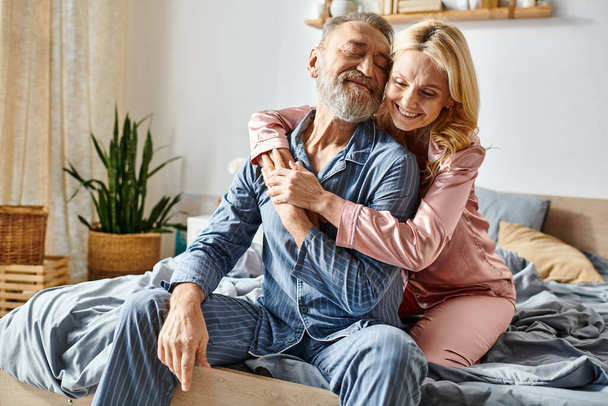 A mature man and woman, in cozy homewear, sit together on a bed, sharing a serene moment of togetherness. - Photo, Image