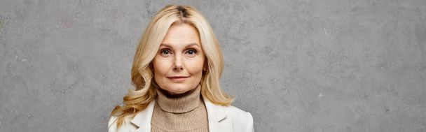 Mature woman exudes sophistication in white jacket and turtle neck sweater against a gray backdrop. - Foto, Imagem