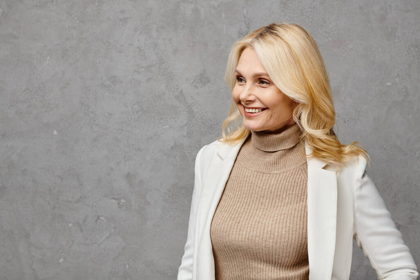 A mature woman exudes elegance in a tan turtleneck sweater and white blazer, striking a pose against a gray backdrop. - Photo, image