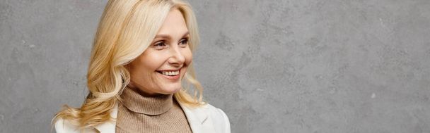 Elegant mature woman with blonde hair and a white jacket smiling charmingly against a gray backdrop. - Fotografie, Obrázek