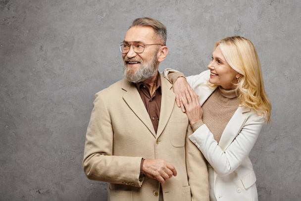 Een volwassen, elegante man en vrouw staan zij aan zij in uitmuntende kleding, ademende liefde en verfijning tegen een grijze achtergrond. - Foto, afbeelding