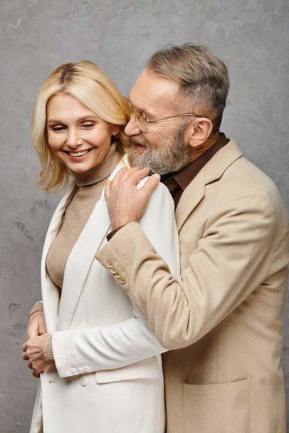 Un hombre y una mujer elegantes y maduros se unen en trajes elegantes sobre un fondo gris. - Foto, Imagen