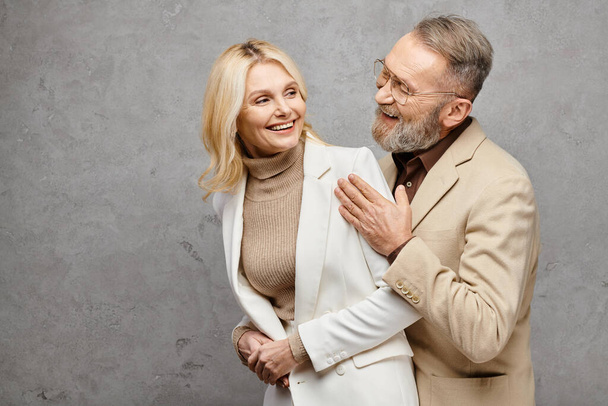 Eleganter, reifer Mann und Frau in Debonair-Kleidung stehen zusammen, verströmen Liebe und Raffinesse vor grauem Hintergrund. - Foto, Bild
