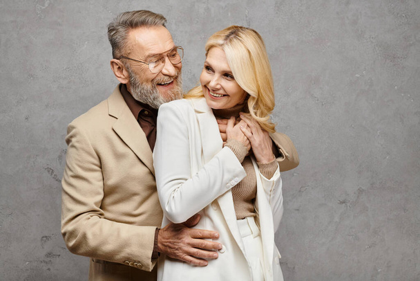 A mature man and woman, elegantly dressed, standing close together in a loving embrace on a gray backdrop. - Photo, Image