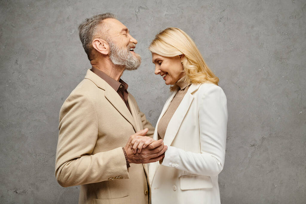 Mature, elegant couple in debonair attire embrace, holding hands lovingly against a gray backdrop. - Foto, imagen